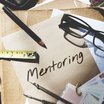 Stifte, Lineale usw. liegen auf einem Schreibtisch. Auf einem Blatt Papier steht "Mentoring" geschrieben