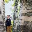 Eine jugendliche Person steht mit einem Fernglas zwischen mehreren Birken und beobachtet die Natur.