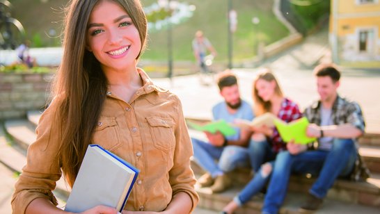 Mädchen hält ein Buch in der Hand, im Hintergrund sind drei Studierende