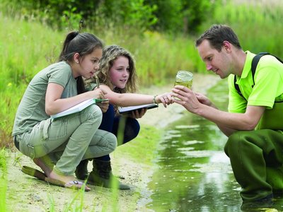 Ein Forscher und zwei Mädchen hocken an einem Gewässer und beobachten Wassertiere in einem Glas.
