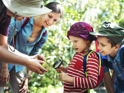 Eine Familie mit zwei kleinen Kindern ist in der Natur unterwegs und erforscht etwas mit einer Lupe.